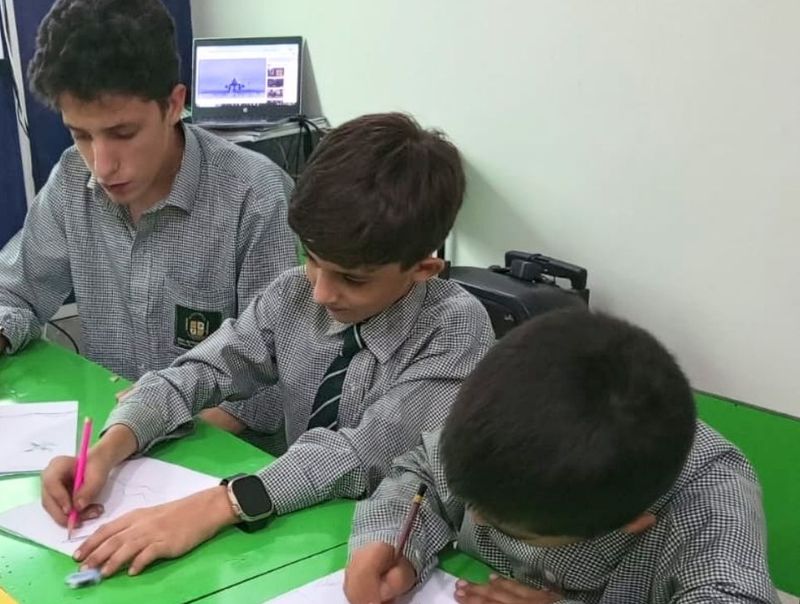Pupils in check uniform drawing at green classroom tables, with a laptop on the desk behind them