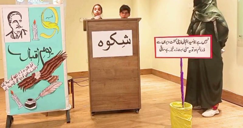 A teacher guiding two pupils as they recite Iqbal's poetry from a lectern on Iqbal Day