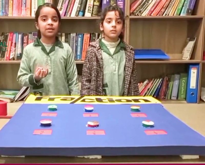 Two pupils presenting a fractions activity board to the class, in front of a bookshelf
