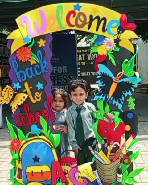 Two young pupils in uniform standing under a colourful Welcome back to school arch at the school entrance