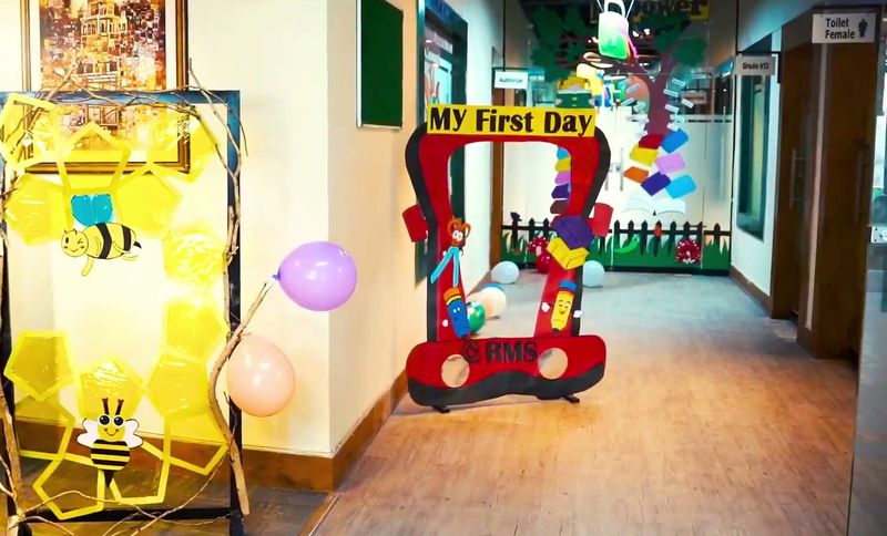 A bright school corridor decorated with a bee-themed door and a My First Day photo frame.
