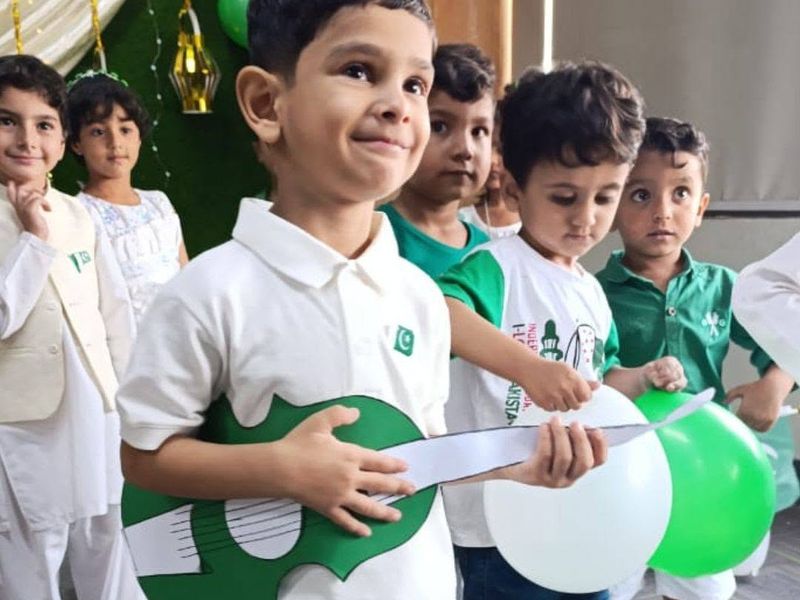 A beaming boy playing a green paper guitar with classmates holding green and white balloons.