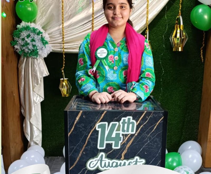 A girl in green and white standing at a 14th August podium decorated with balloons and lanterns.