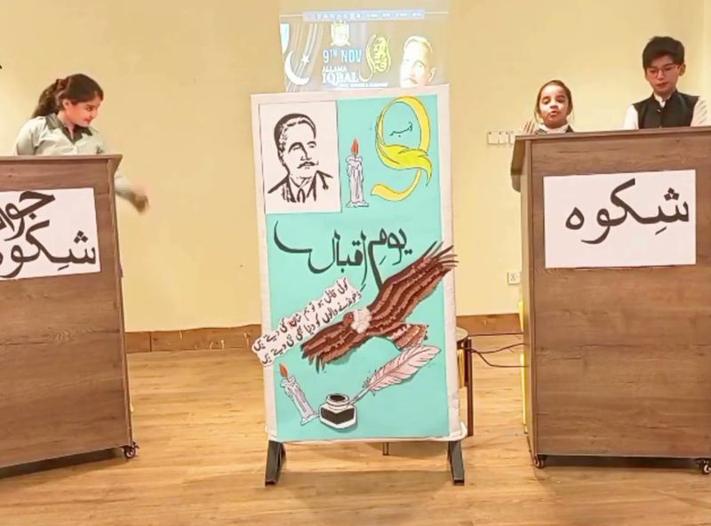 Pupils at two lecterns reciting Iqbal's Shikwa and Jawab-e-Shikwa beside an Iqbal Day display.