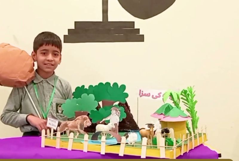 A boy presenting a handmade model of an Urdu moral story with a shepherd, sheep and a lion.