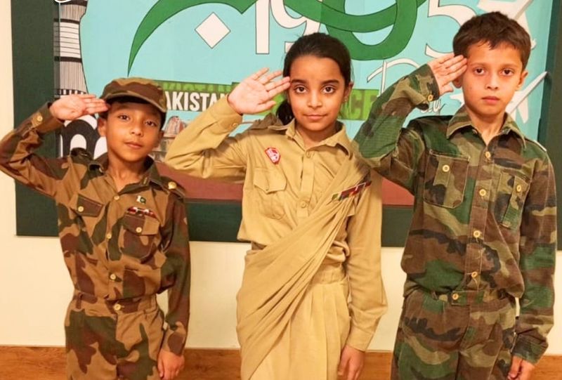 Three pupils in army and nurse costumes saluting in front of the 6 September 1965 Defence Day backdrop.