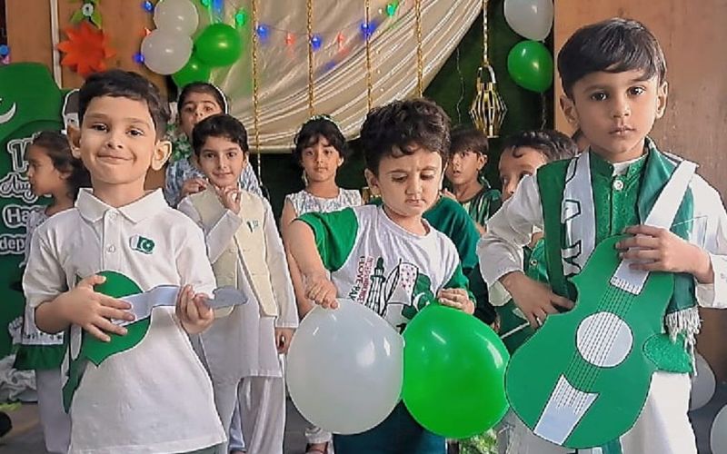 Young pupils in green and white outfits with balloons and paper guitars at the Independence Day celebration.