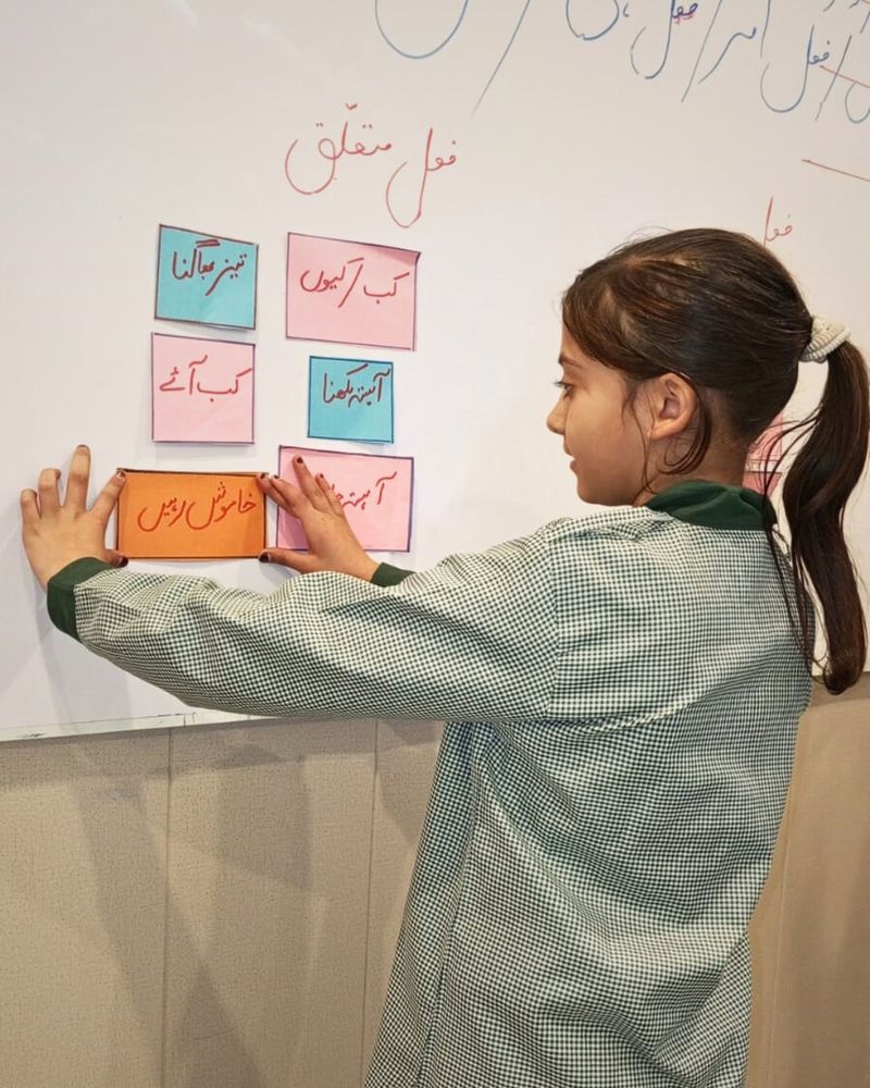 A pupil in the RMS check smock placing Urdu grammar word cards on the classroom whiteboard.
