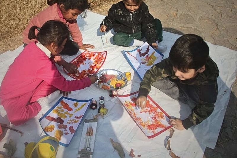 Early Years children sitting on a sheet outdoors, painting autumn leaf pictures with brushes and real leaves.
