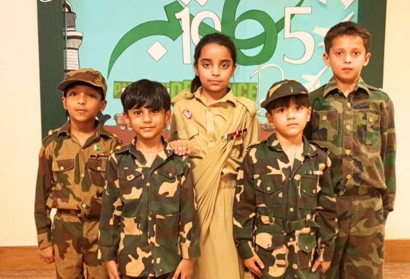 Five pupils in army camouflage and a khaki uniform standing in front of a 6 September 1965 Defence Day backdrop.