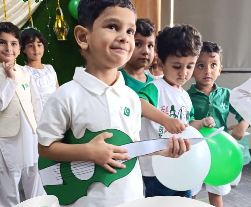 A smiling young pupil in a white shirt holding a green paper guitar, with classmates and green and white balloons behind.
