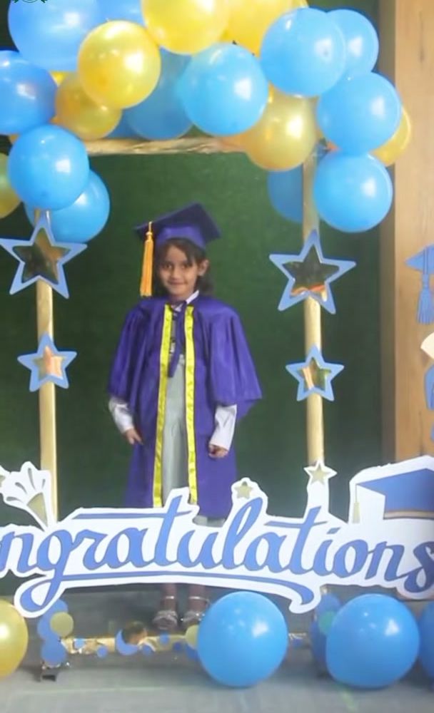 A kindergarten graduate in a purple gown and cap standing under a blue and gold balloon arch with a Congratulations sign.