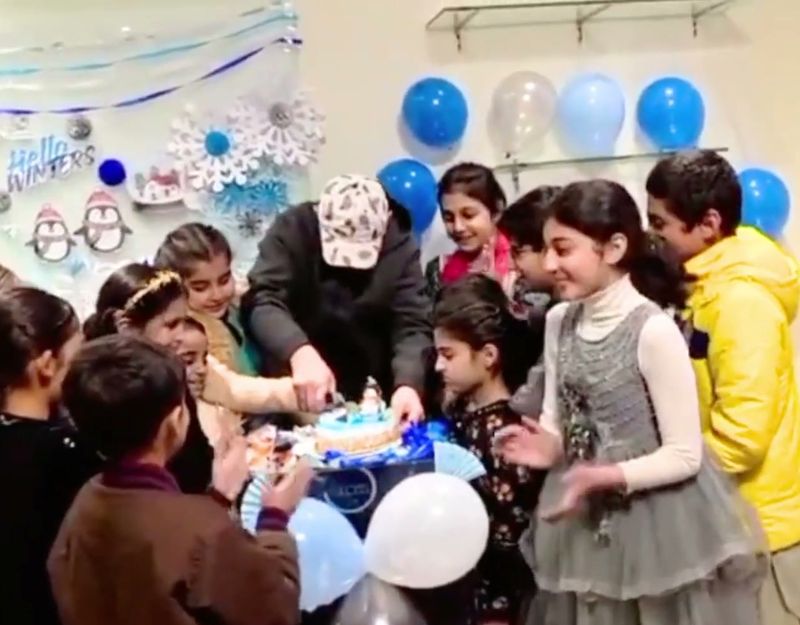 Primary pupils in party clothes gathered around a table to cut a winter-themed cake, with blue and white balloons behind.