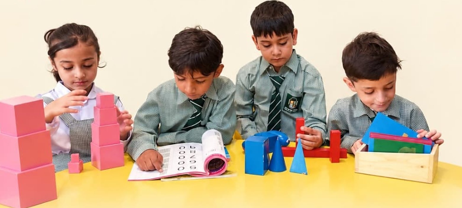Four RMS students in green check uniforms working with Montessori materials: the pink tower, a number book, geometric solids and a book box.